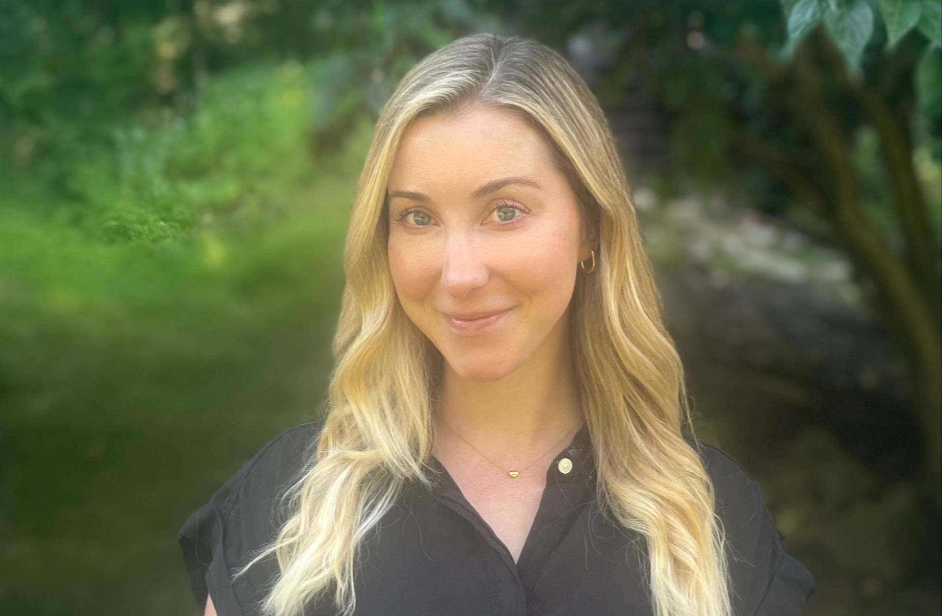 A woman with long, wavy blonde hair wearing a black top and a gold necklace smiles softly while standing outdoors with greenery blurred in the background.