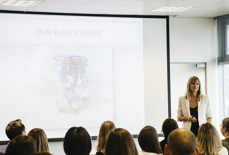 A woman stands in front of a seated audience, presenting a slideshow. The projected slide reads OUR YEARLY WASTE and shows a jar filled with waste. The room is bright with natural light from large windows.