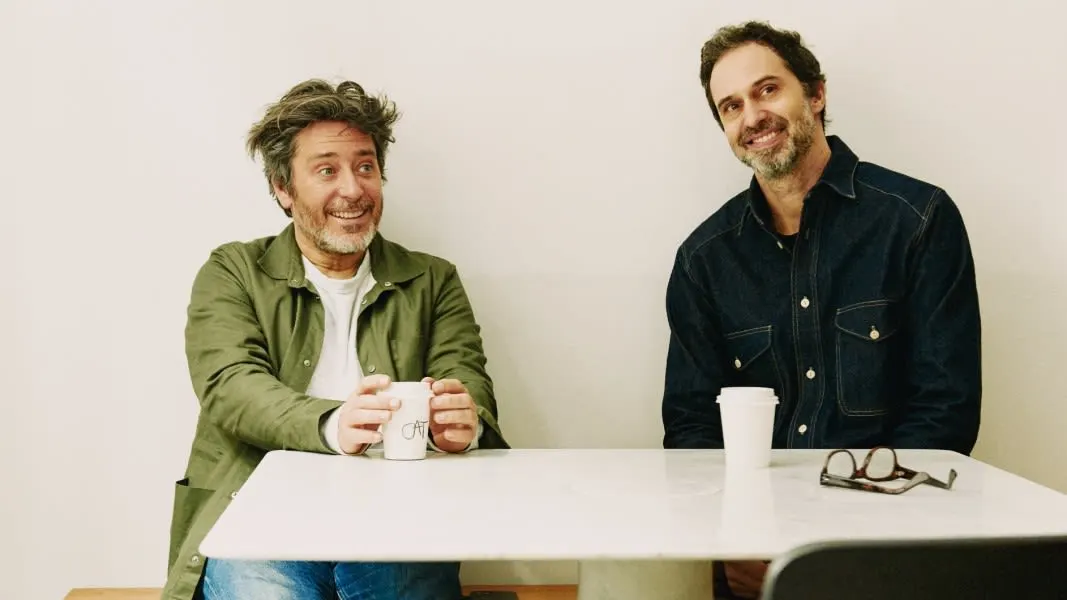 Two men sit at a marble table against a white wall. One smiles widely holding a mug, while the other sits with a relaxed smile and a white takeaway cup. A pair of eyeglasses is on the table.