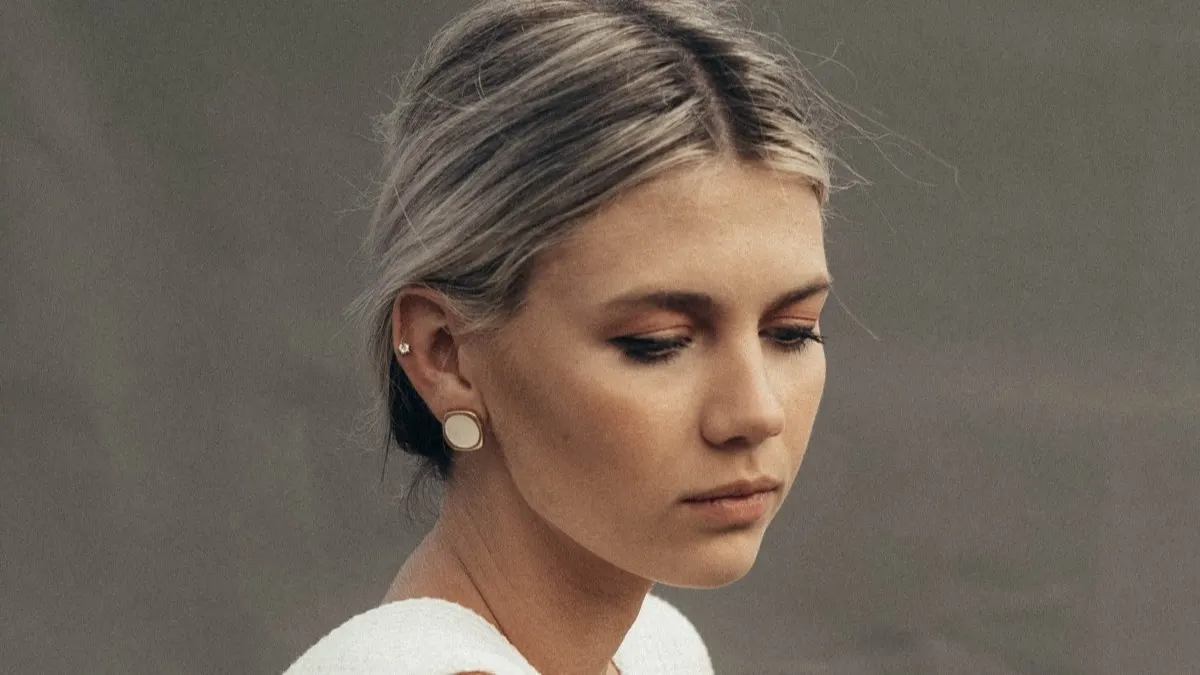 A young woman with light hair tied back looks downward with a neutral expression. She wears a white top and round earrings. The background is plain and muted.