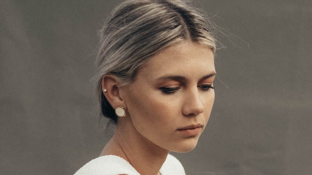 A young woman with light hair tied back looks downward with a neutral expression. She wears a white top and round earrings. The background is plain and muted.