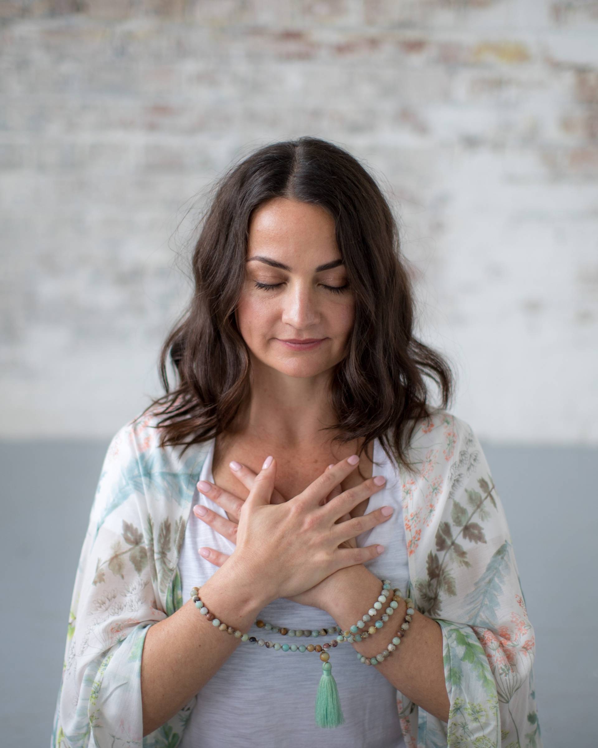 A woman with long brown hair stands with her eyes closed, hands crossed over her chest. She wears a light floral shawl, a gray top, and beaded bracelets, appearing calm and peaceful in a softly lit room with a faded brick wall.
