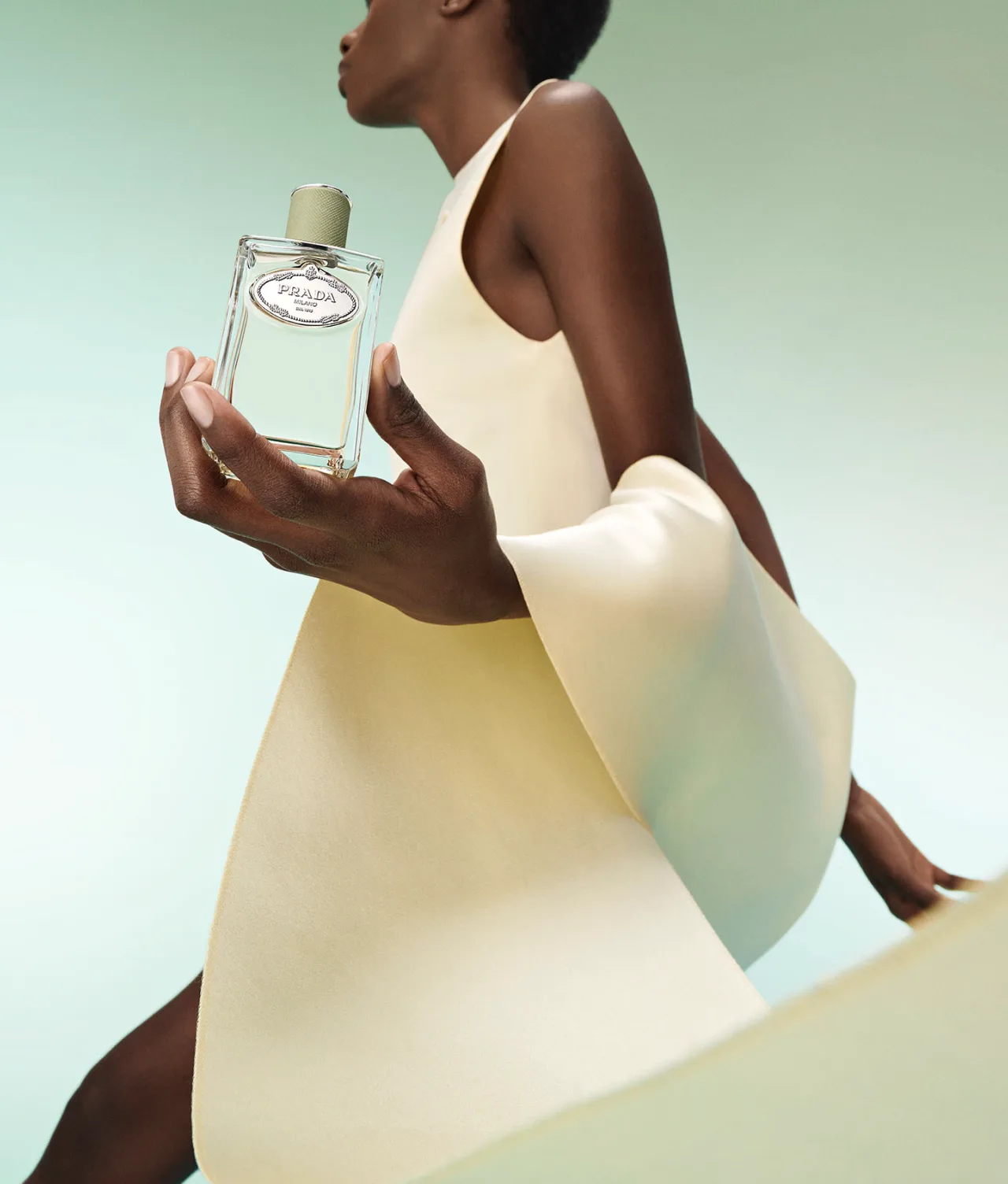 A person in an elegant, cream-colored outfit holds a bottle of Prada perfume against a soft green background, with their arm gracefully extended and the perfume bottle prominently displayed.
