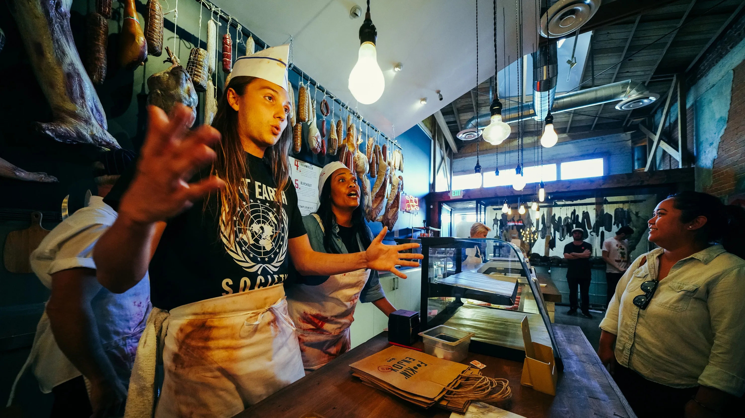 Two people wearing aprons and hats animatedly talk to a customer in a rustic deli with hanging meats, a display counter, and warm lighting. Another person listens from across the counter.