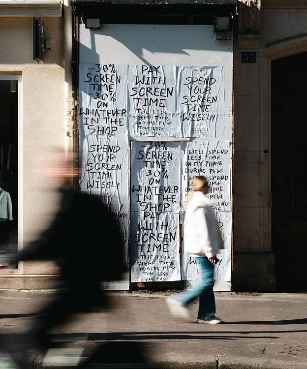 A person walks past a storefront covered with white posters that read “Pay with screen time. 30% on whatever in the shop. Spend your screen time wisely” in black handwritten letters. Another blurred person walks by.
