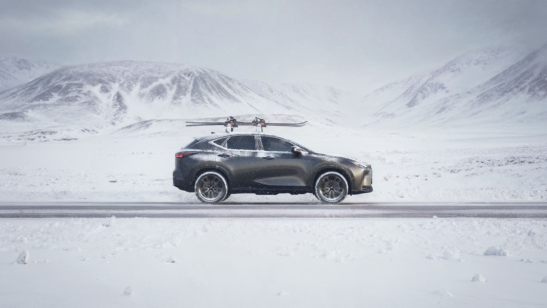 A dark SUV with a roof rack drives on a snowy road, surrounded by snow-covered mountains under a cloudy sky.