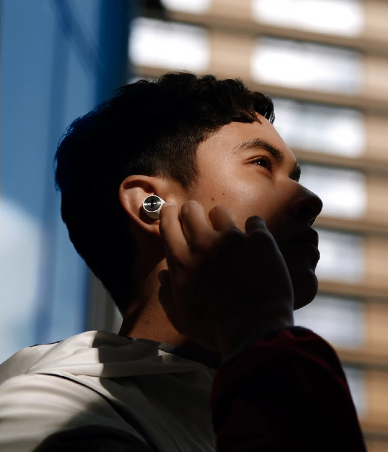 A person in a white jacket stands outside near a building, inserting a wireless earbud into their ear, with sunlight highlighting part of their face.