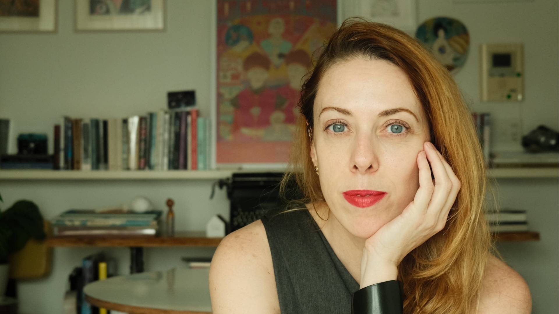 A woman with long, light brown hair and red lipstick rests her face on her hand, looking at the camera. She sits indoors in front of shelves with books, framed art, and a round table.