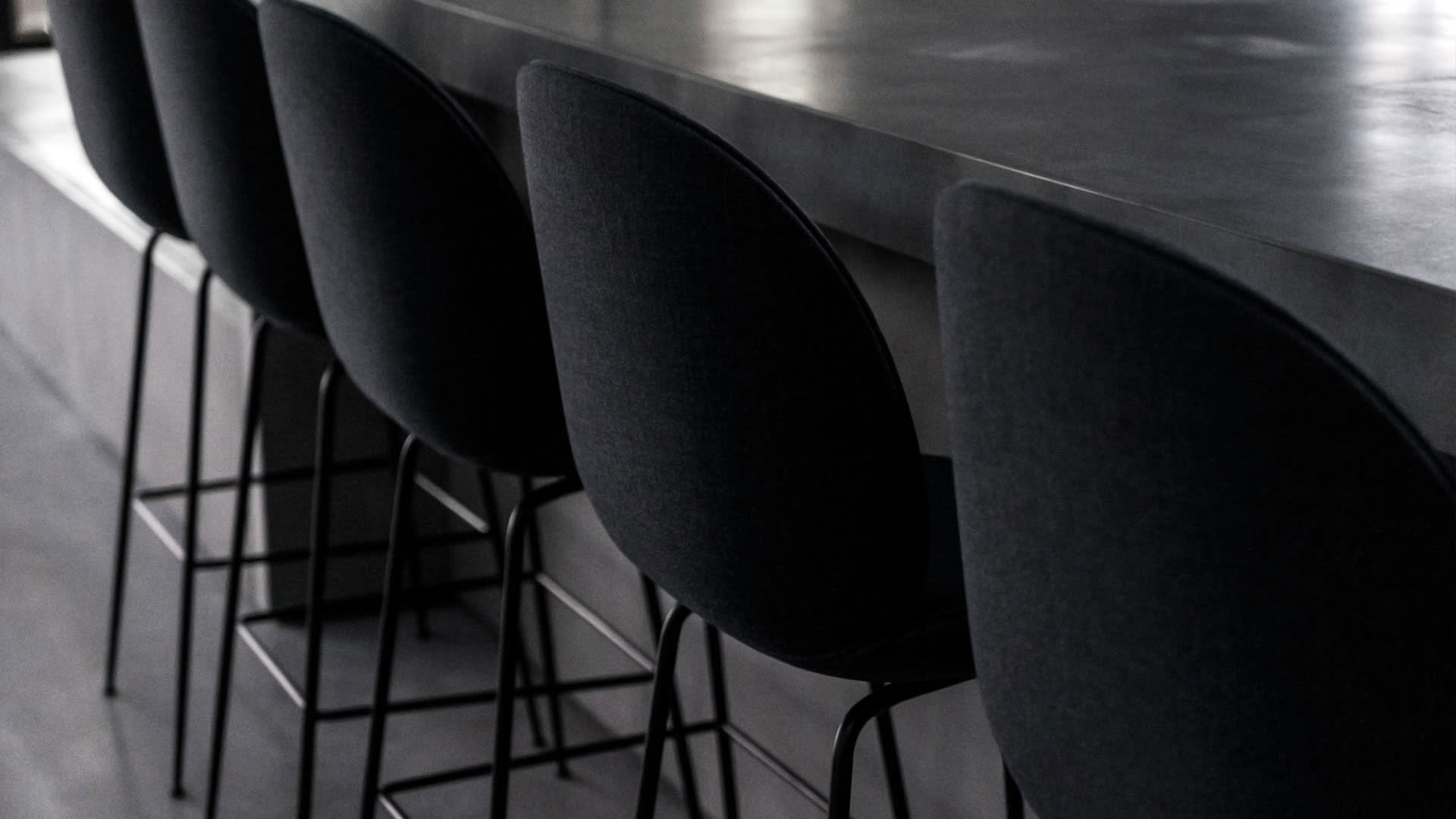 Four modern black barstools with curved backs are lined up along a sleek, dark countertop in a minimalist, contemporary setting. The lighting is dim, creating a stylish and sophisticated atmosphere.