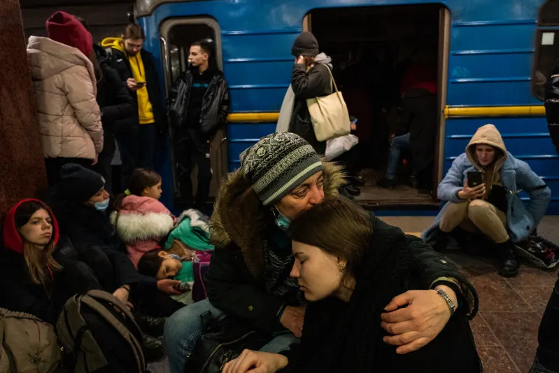 People sit on the floor of a subway station next to a blue train. A woman wearing a knit hat hugs and comforts a young girl, while others sit, stand, or use their phones around them. The atmosphere appears tense and somber.