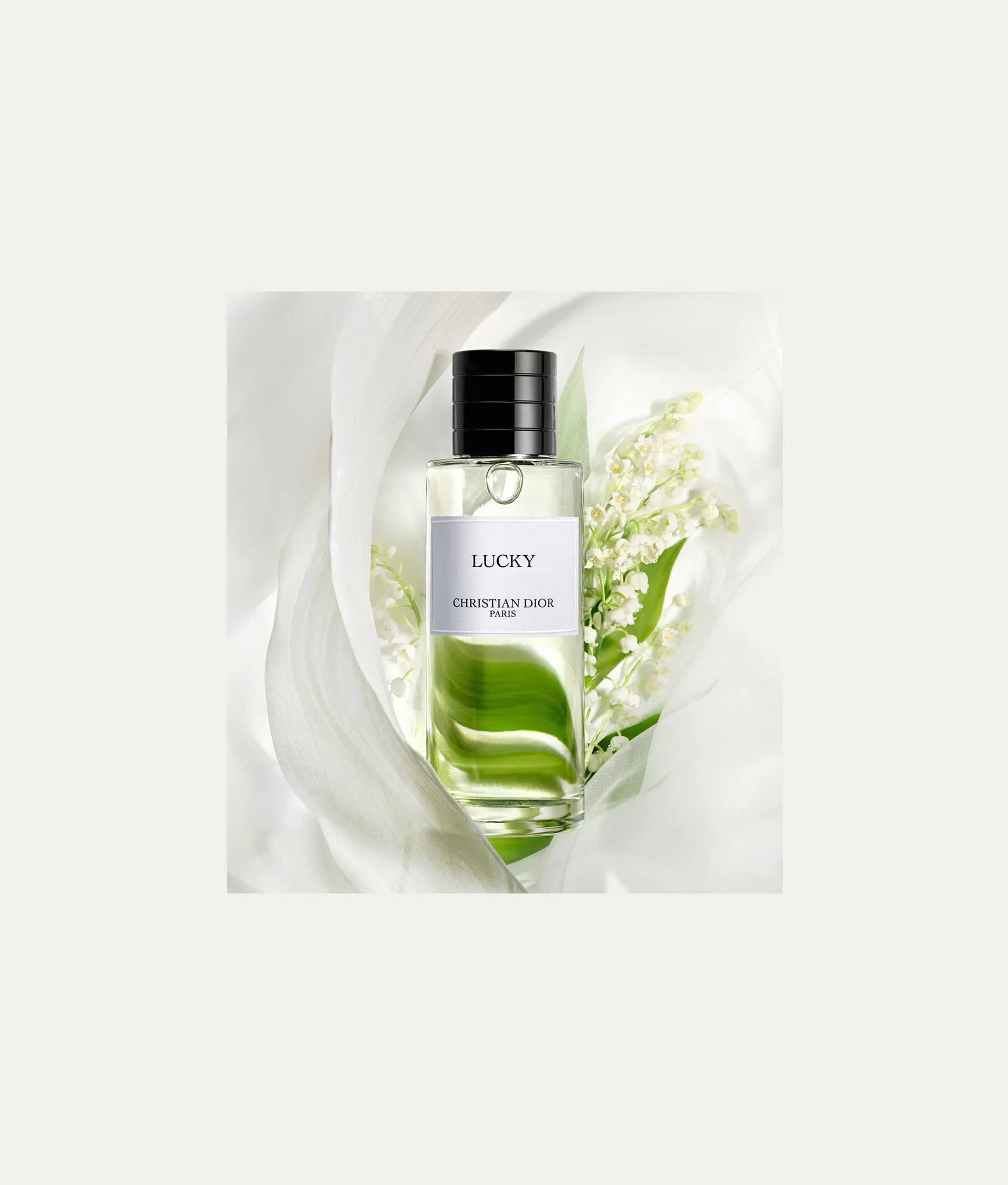 A clear bottle of Dior Lucky perfume with a black cap is surrounded by white fabric and delicate lily of the valley flowers, creating a soft and elegant composition.
