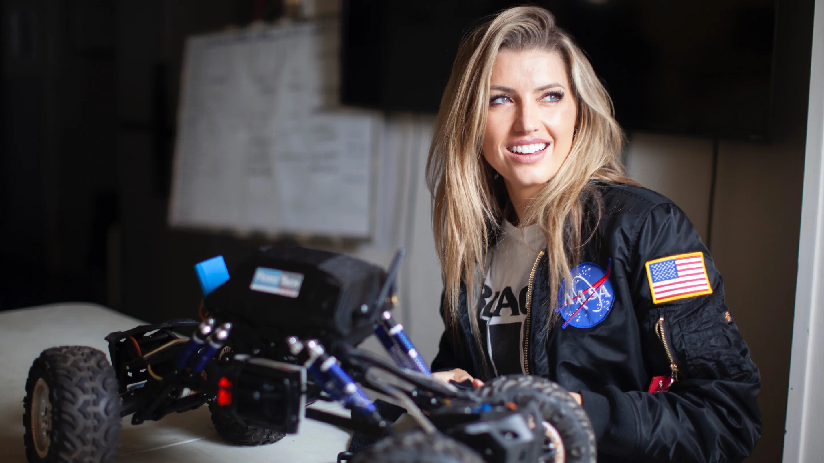 A woman wearing a NASA jacket smiles while seated at a table with a partially assembled remote-controlled vehicle or robot in front of her. The background is softly blurred.