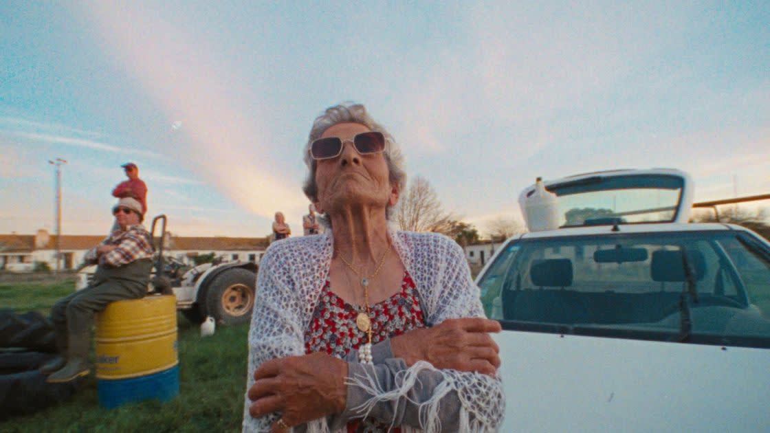 An elderly woman wearing sunglasses, a white shawl, and floral dress stands with arms crossed in front of a white car, while people and a tractor are seen in the background under a blue sky.