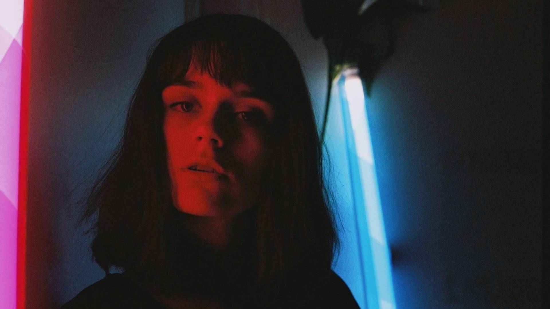 A woman with shoulder-length dark hair stands in moody lighting, her face illuminated by red and blue neon lights behind her, creating a dramatic and artistic contrast.