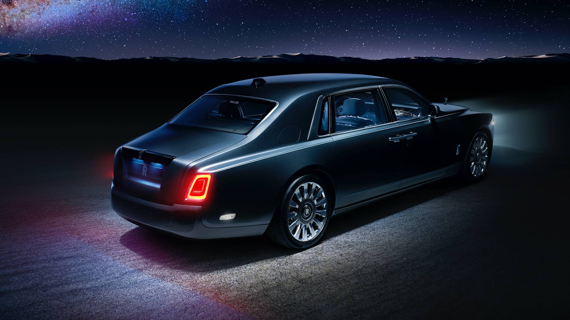 A luxury black sedan with illuminated taillights is parked on a road at night, with mountains and a starry sky in the background.