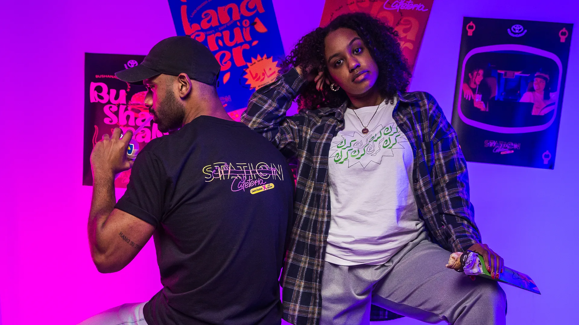 Two people pose confidently in front of colorful posters. One, in a black cap and t-shirt with a bold graphic, sits backward, while the other, with curly hair and a plaid shirt, leans beside him wearing a white graphic tee. Purple-blue lighting.