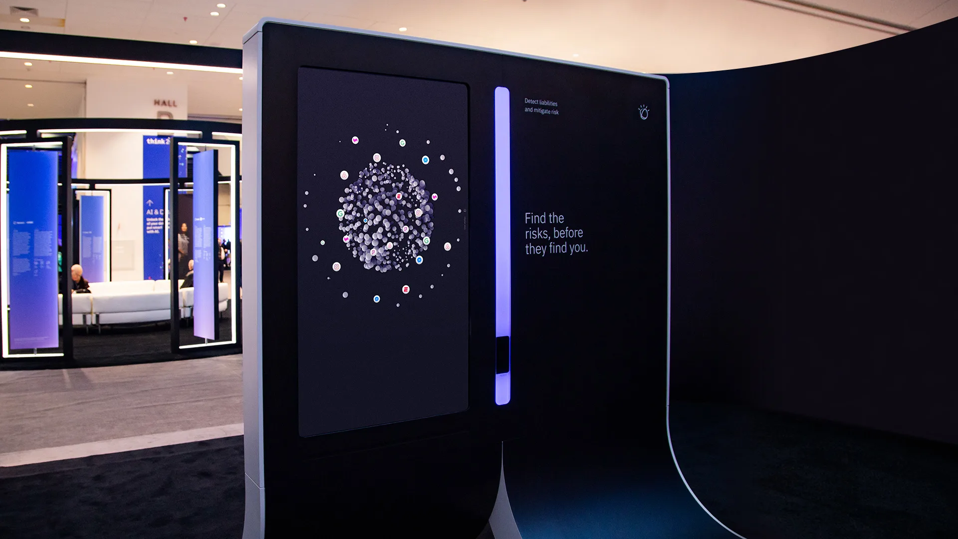 A digital display at an indoor exhibit shows a clustered data visualization. Text reads, Find the risks, before they find you. The modern booth features minimalist design and glowing blue accents.
