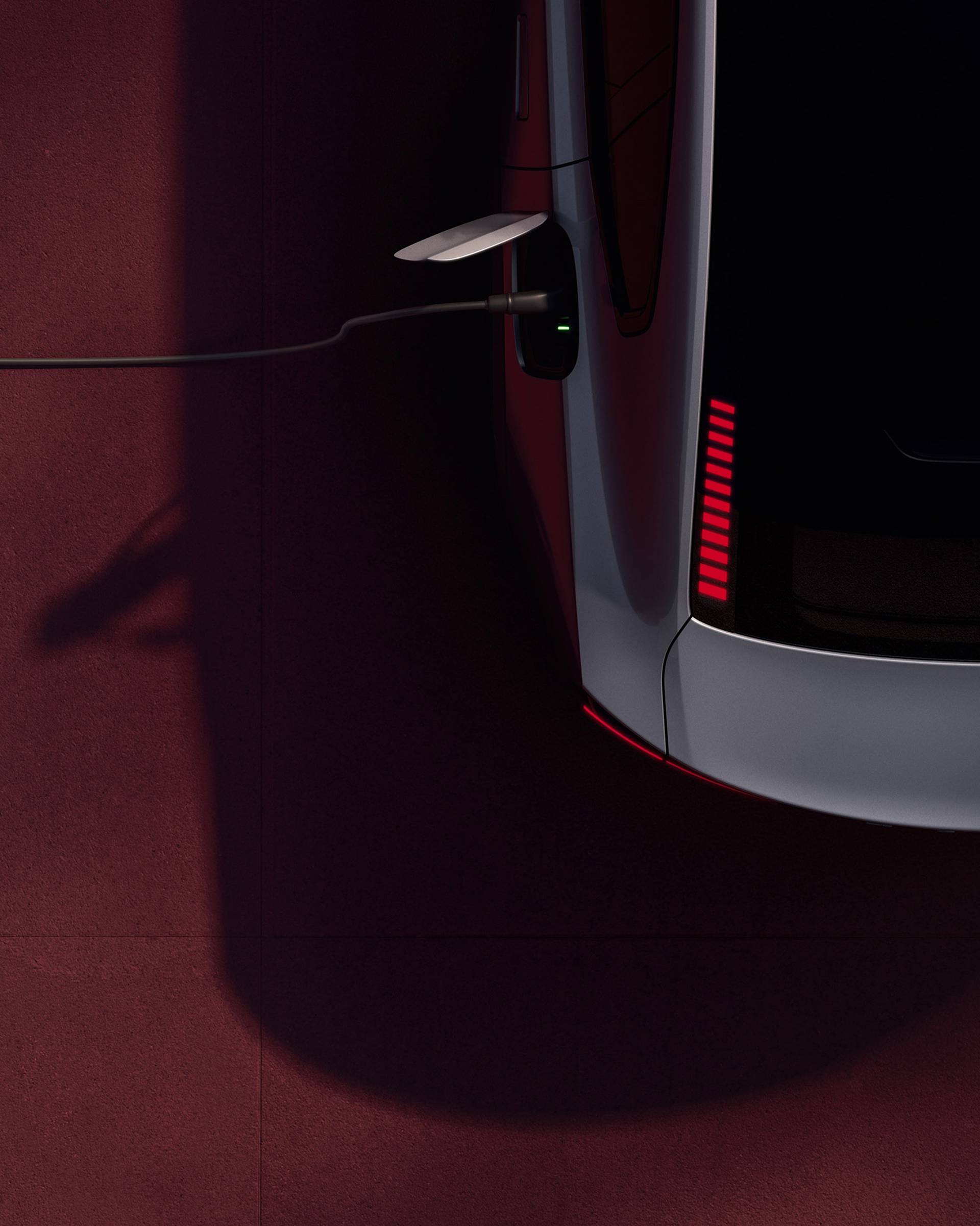 A top-down view of a silver electric car charging on a red surface, with the charging cable plugged into the vehicle and a line of red brake lights visible on the back.