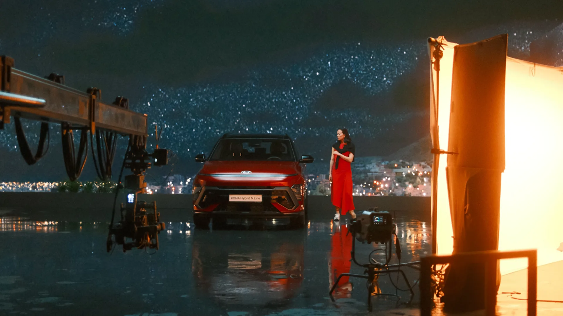 A woman in a red dress stands beside a red Volkswagen Concept car on a wet reflective surface, with studio lights, cameras, and equipment visible around her. A starry night backdrop is displayed behind the scene.