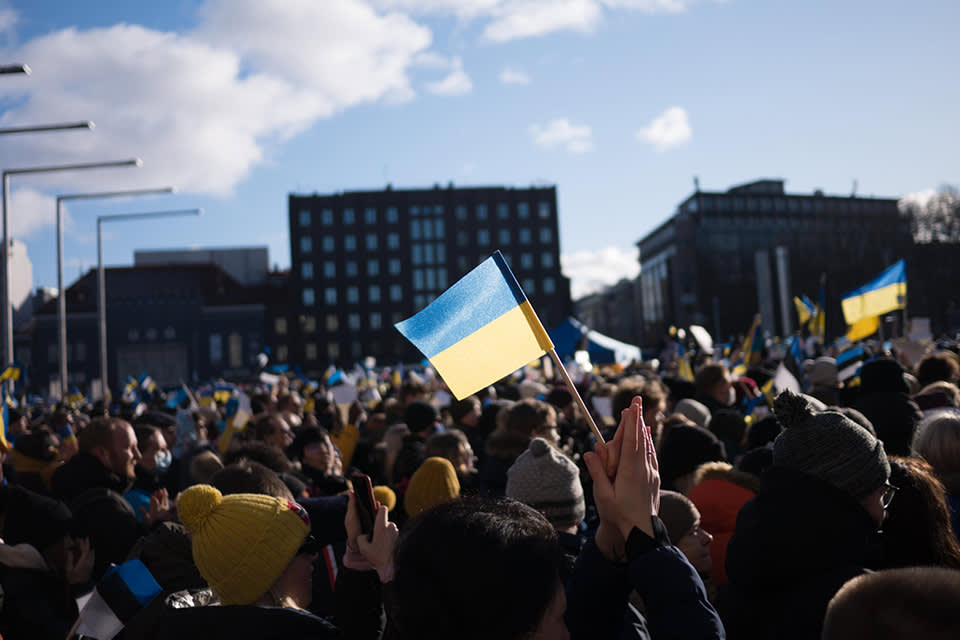 A large crowd gathers outdoors, many holding Ukrainian flags. People are dressed in winter clothing, and the background shows several buildings under a sunny sky.