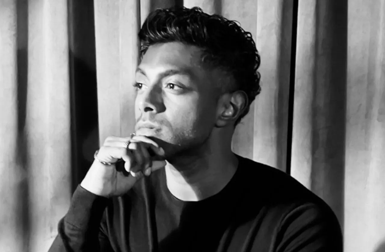 A man with short, curly hair sits in front of curtains, resting his chin on his hand and looking thoughtfully into the distance. The image is in black and white.