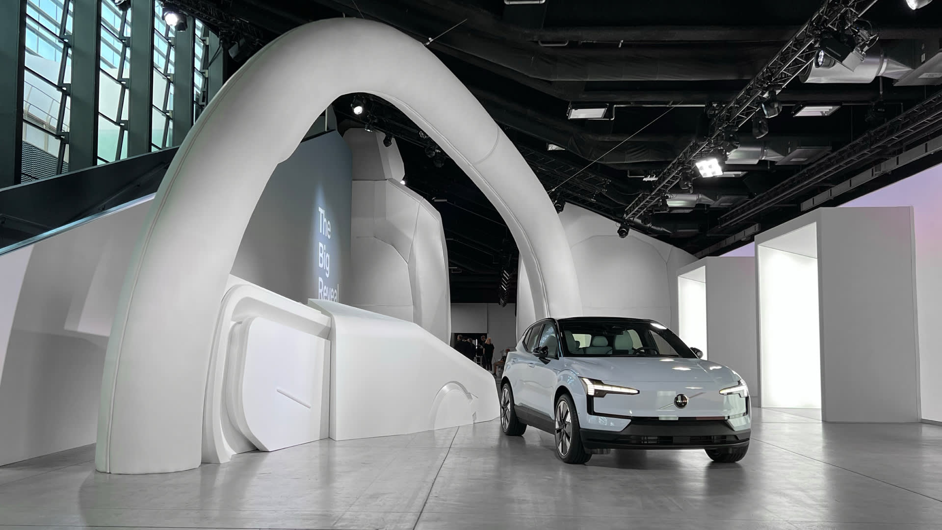 A white electric car is displayed in a modern, futuristic showroom with curved white walls and an arch structure under bright lighting.