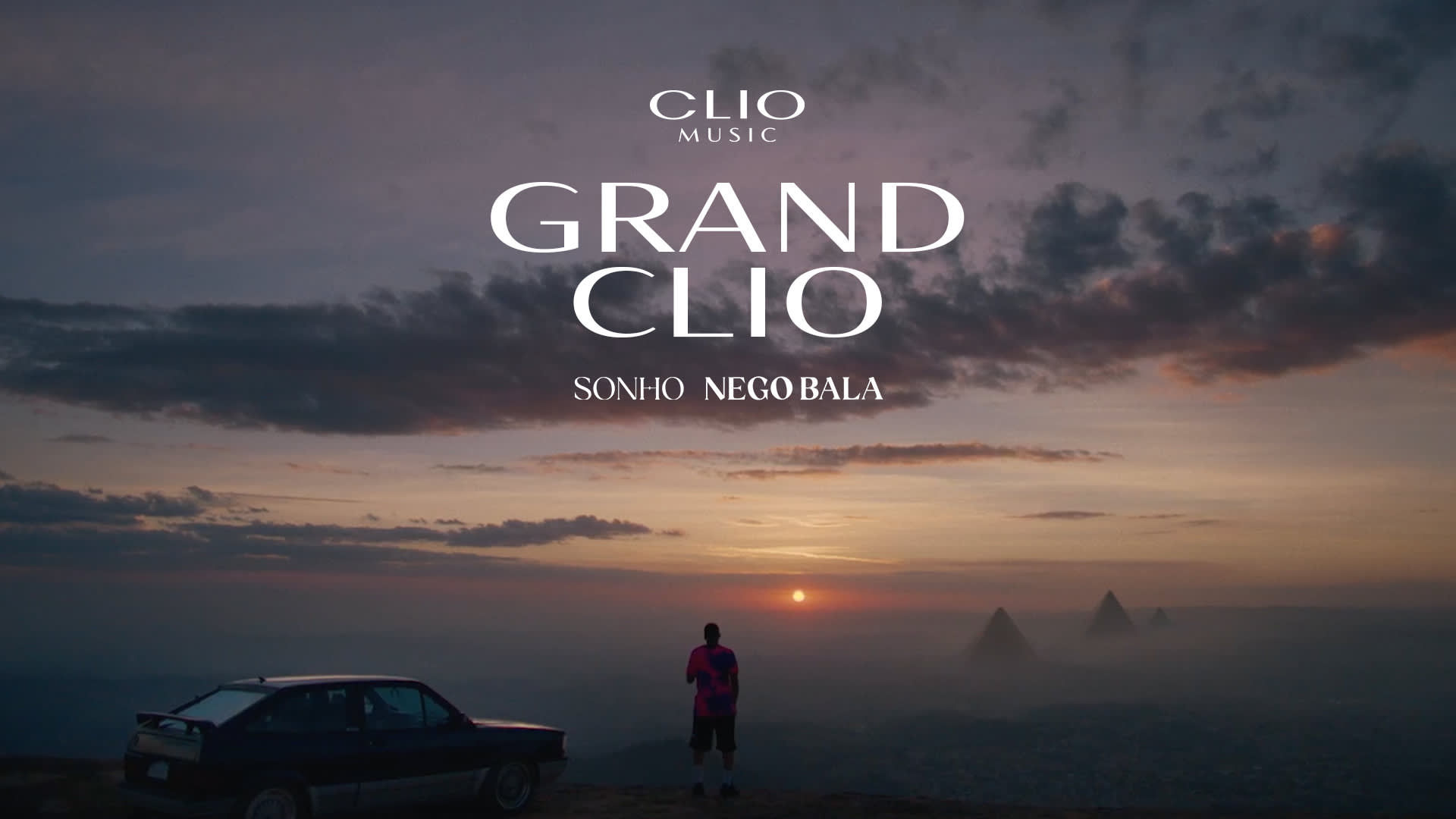 A person stands next to a car on a hilltop at sunset, overlooking a hazy cityscape with pyramids in the distance. Text overlays read CLIO MUSIC GRAND CLIO SONHO NEGO BALA.