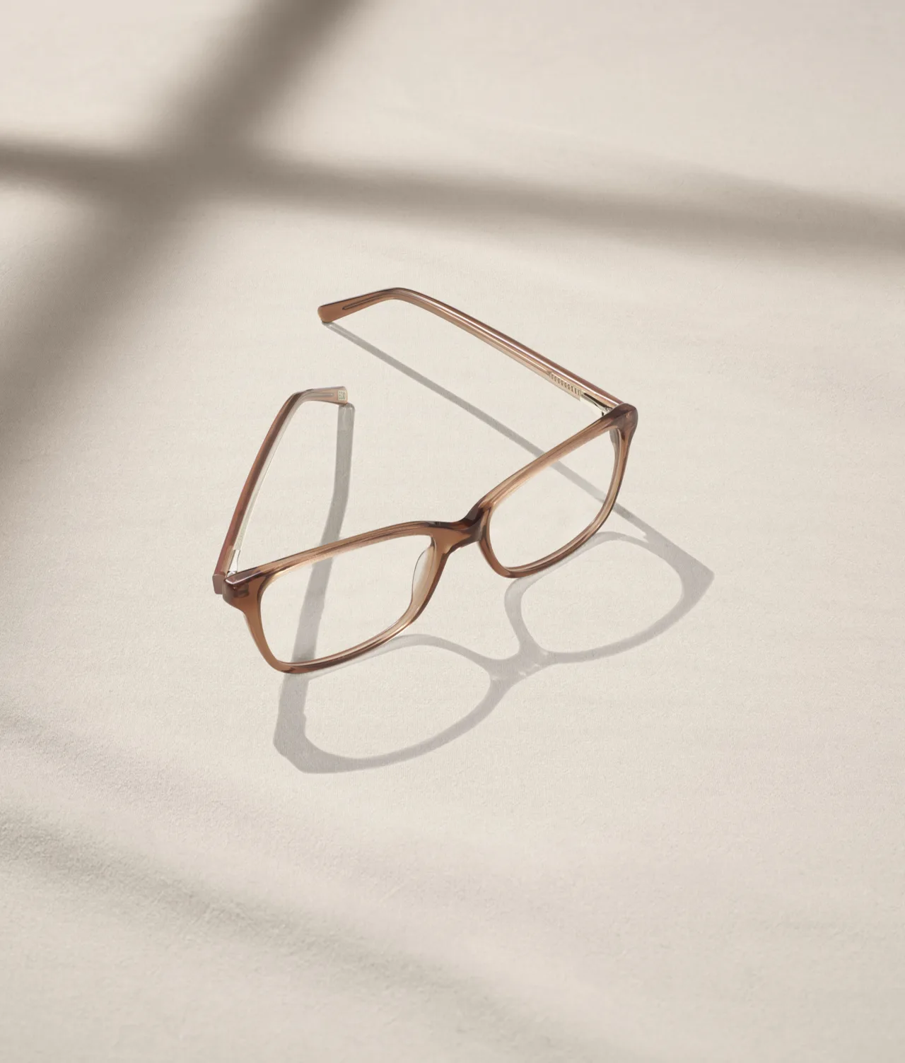 A pair of brown eyeglasses sits on a light beige surface, casting a distinct shadow. Sunlight streams in, creating soft, geometric patterns across the background.