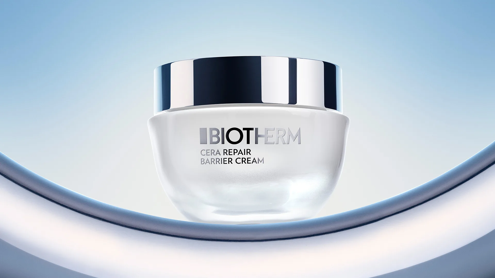 A jar of Biotherm Cera Repair Barrier Cream with a silver lid is displayed against a gradient blue background, positioned on a curved, shiny metallic surface.