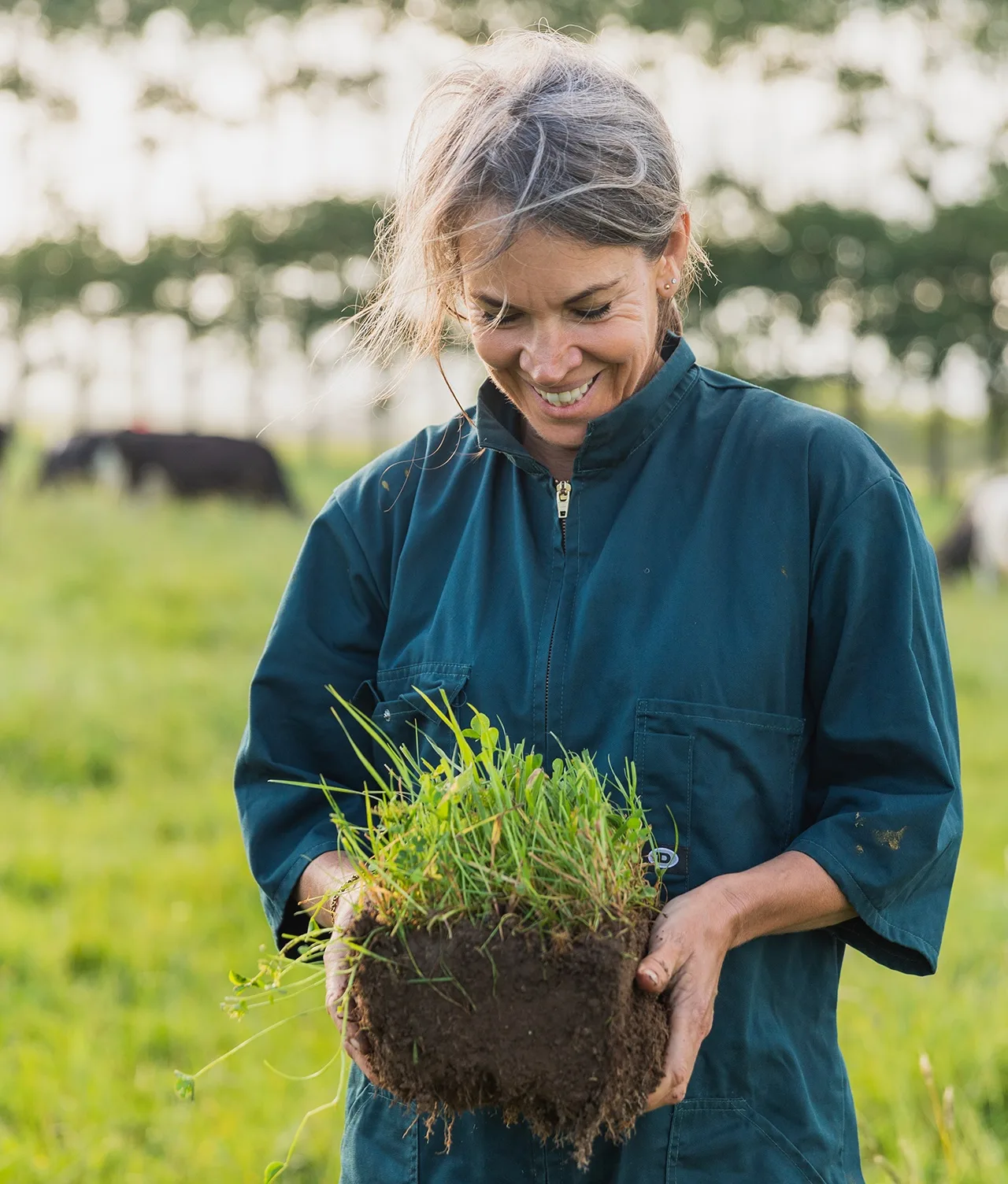 A smiling woman in a blue jumpsuit holds a large clump of soil with grass and roots in a green field, with blurred cows and trees in the background.
