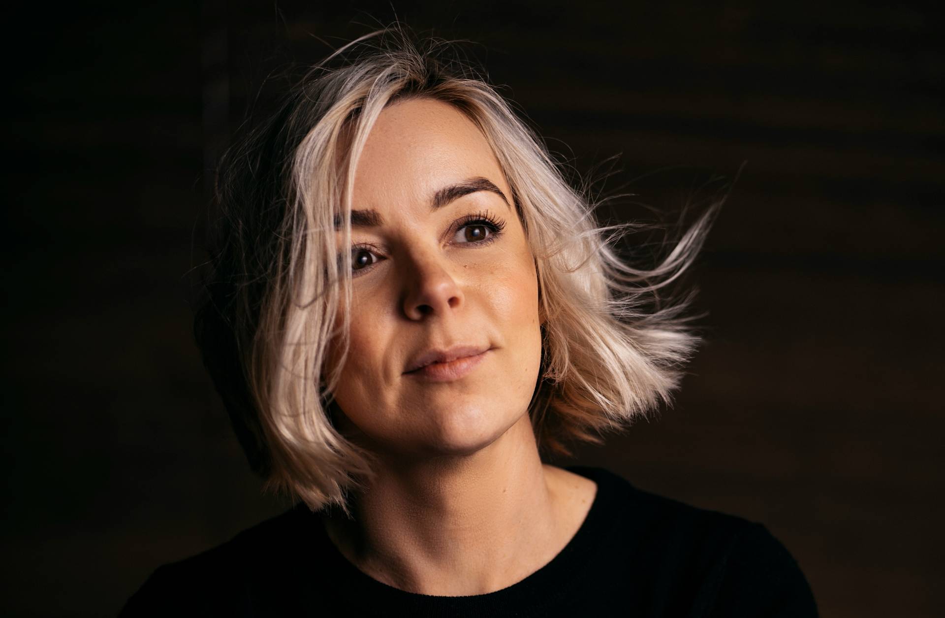 A woman with short, blonde hair and a black top looks slightly upward, softly lit against a dark, blurred background. Her expression is calm and contemplative.