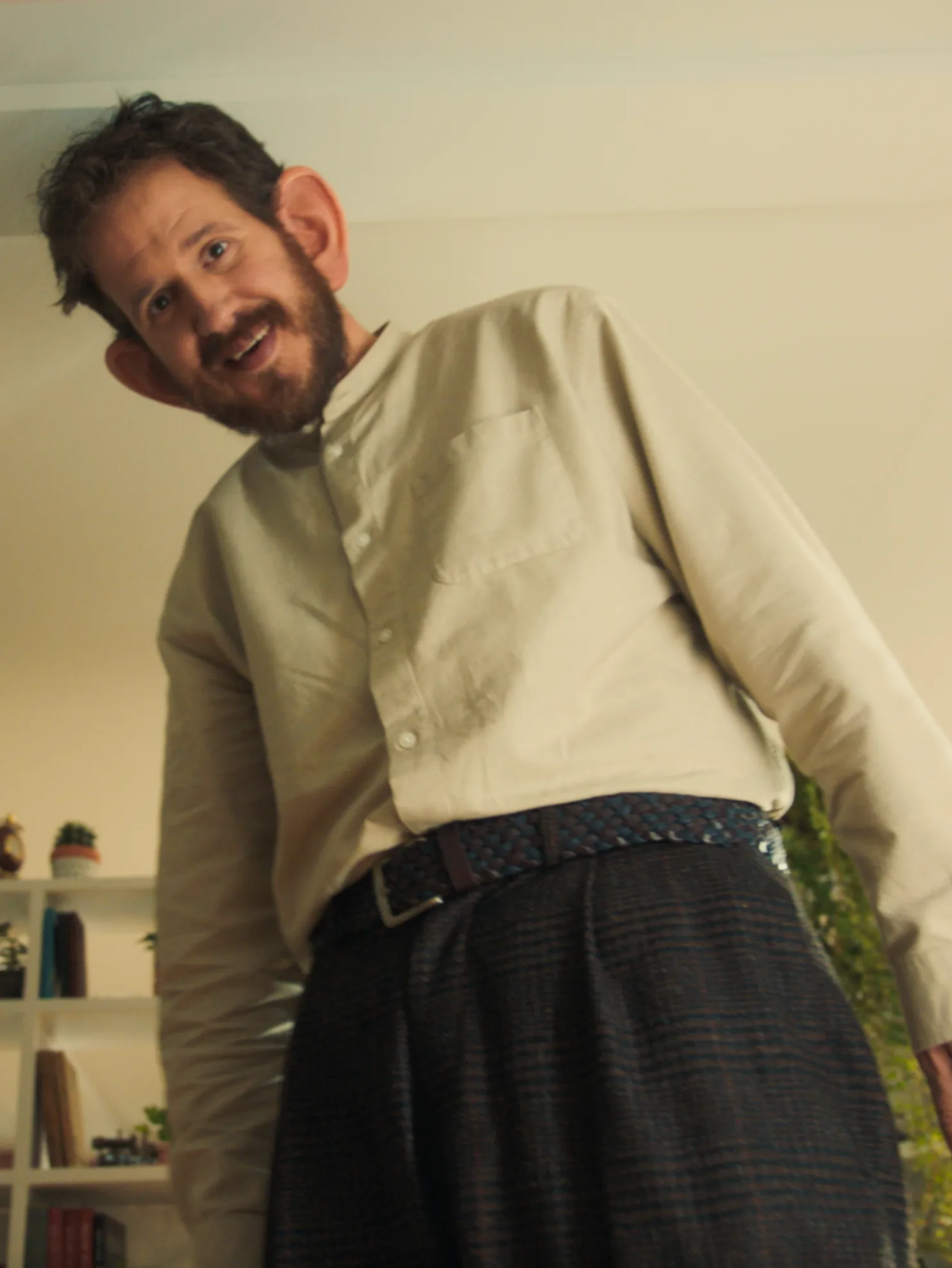 A man with an exaggeratedly large head and ears, wearing a beige shirt and dark pants, smiles while leaning forward in a brightly lit room with shelves and plants in the background.