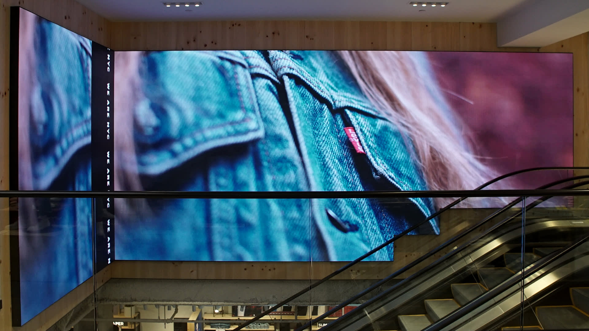A large digital display in a store shows a close-up of a denim jacket with a visible red tag, positioned above an escalator. Part of the image extends onto an adjacent screen on the left.