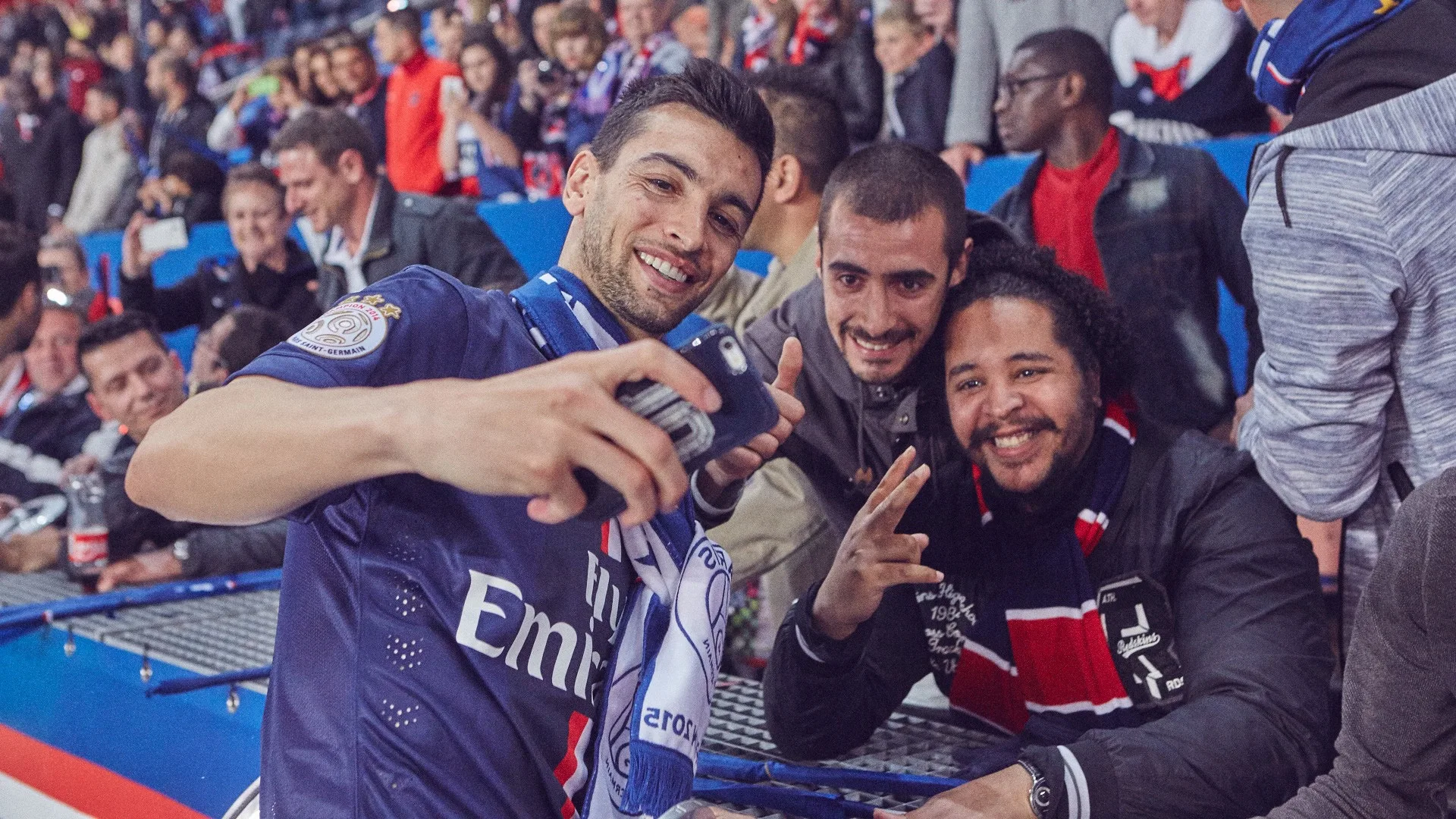 A soccer fan wearing a Paris Saint-Germain jersey takes a selfie with two friends in a crowded stadium. Smiling and making peace signs, they are surrounded by other cheering fans in the stands.