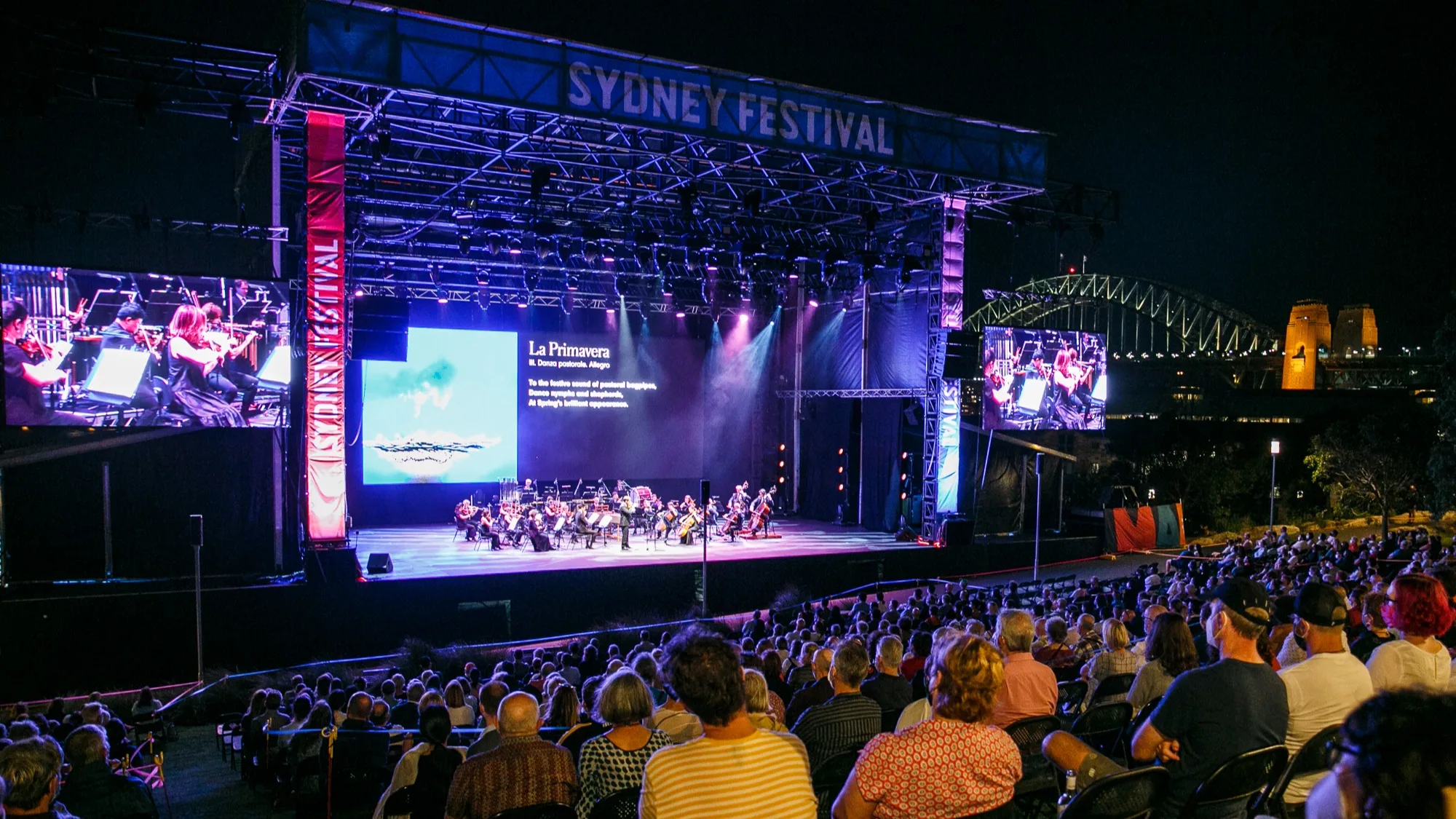 A large audience watches an outdoor orchestral performance on a lit stage at night during the Sydney Festival, with the Sydney Harbour Bridge visible in the background. Two large screens display the event to the crowd.
