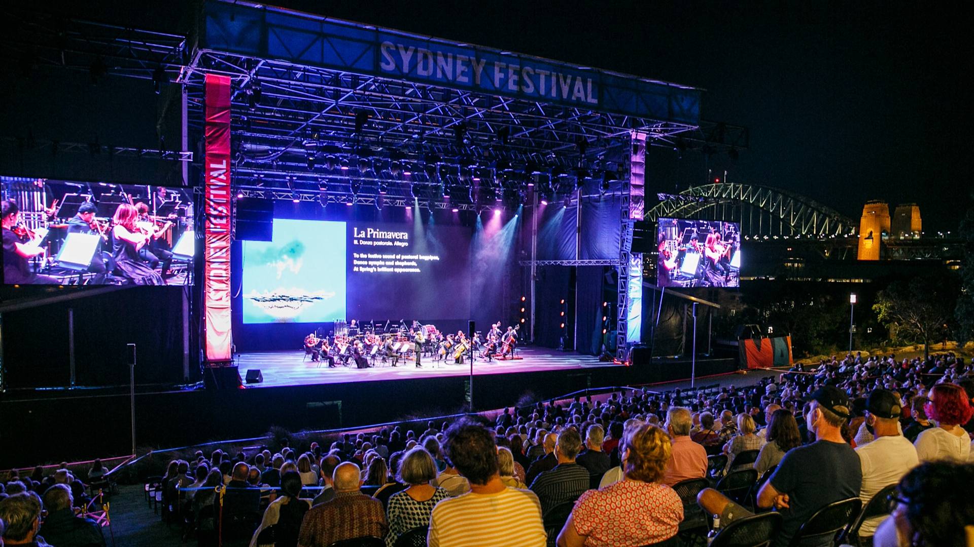 A large audience watches an outdoor orchestral performance on a lit stage at night during the Sydney Festival, with the Sydney Harbour Bridge visible in the background. Two large screens display the event to the crowd.