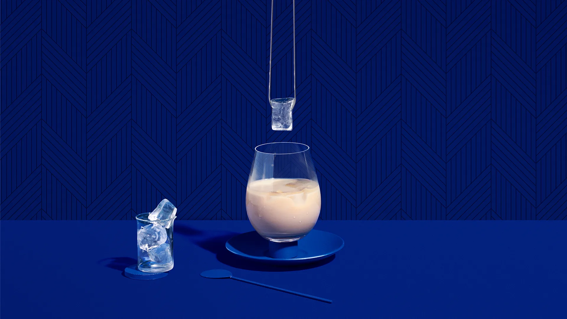 A glass of creamy beverage with ice sits on a blue saucer next to a glass of ice cubes and a blue spoon, all set against a patterned blue background. An ice cube is suspended above the main glass.