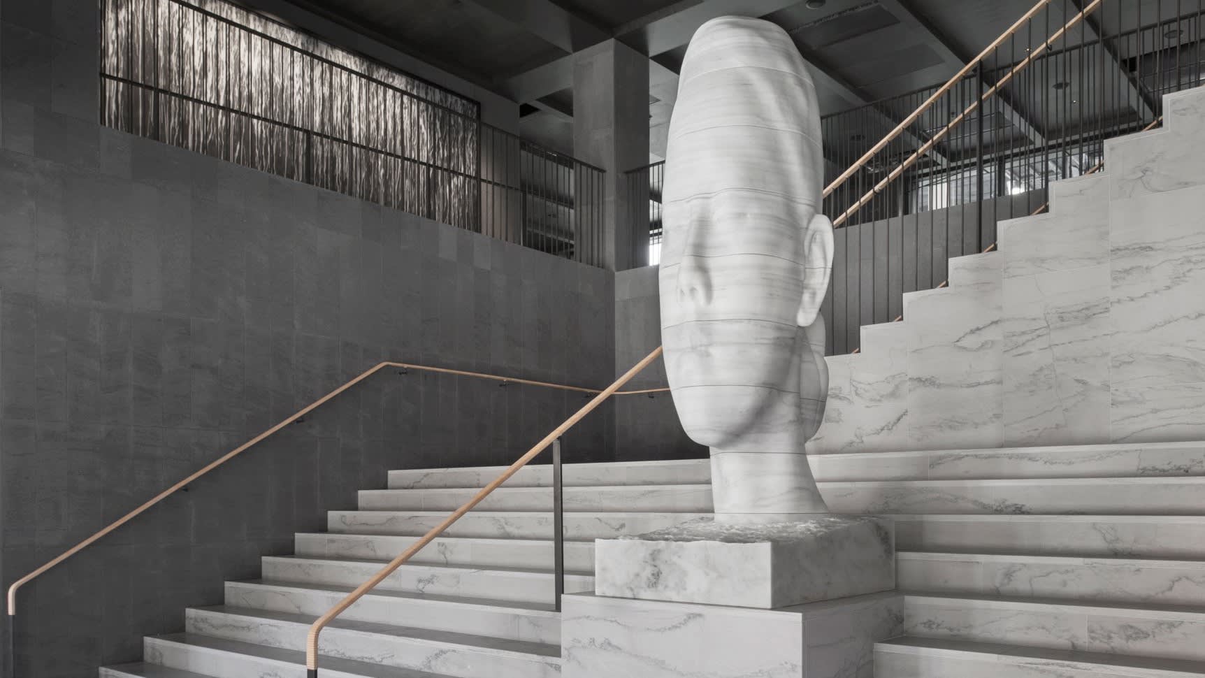 A large, elongated marble sculpture of a human head stands on a staircase in a modern, minimalist interior with gray stone walls, metal railings, and clean lines.