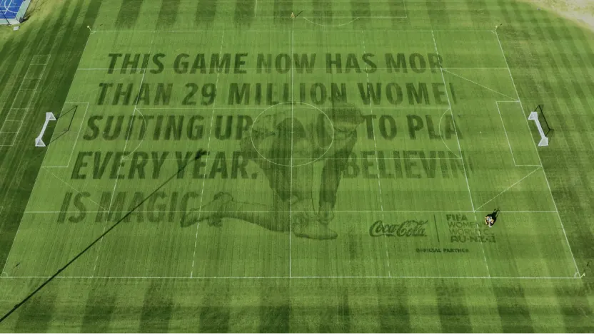 A soccer field with large text on the grass reads, This game now has more than 29 million women suiting up to play every year. Believ is magic. Coca-Cola and FIFA Women’s World Cup logos are visible below.