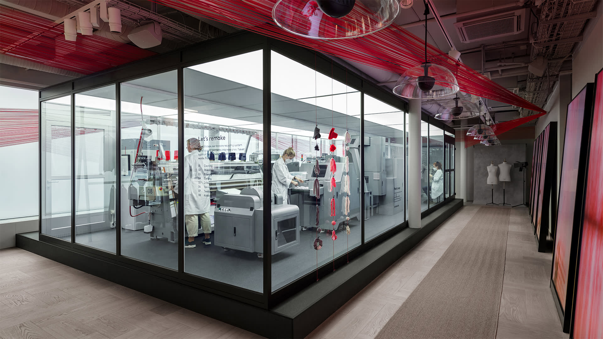 Two people in white coats work with machines in a modern glass-walled lab. Red threads stretch across the ceiling, and mannequins stand in the hallway outside the lab. The space is bright and clean, with a contemporary design.