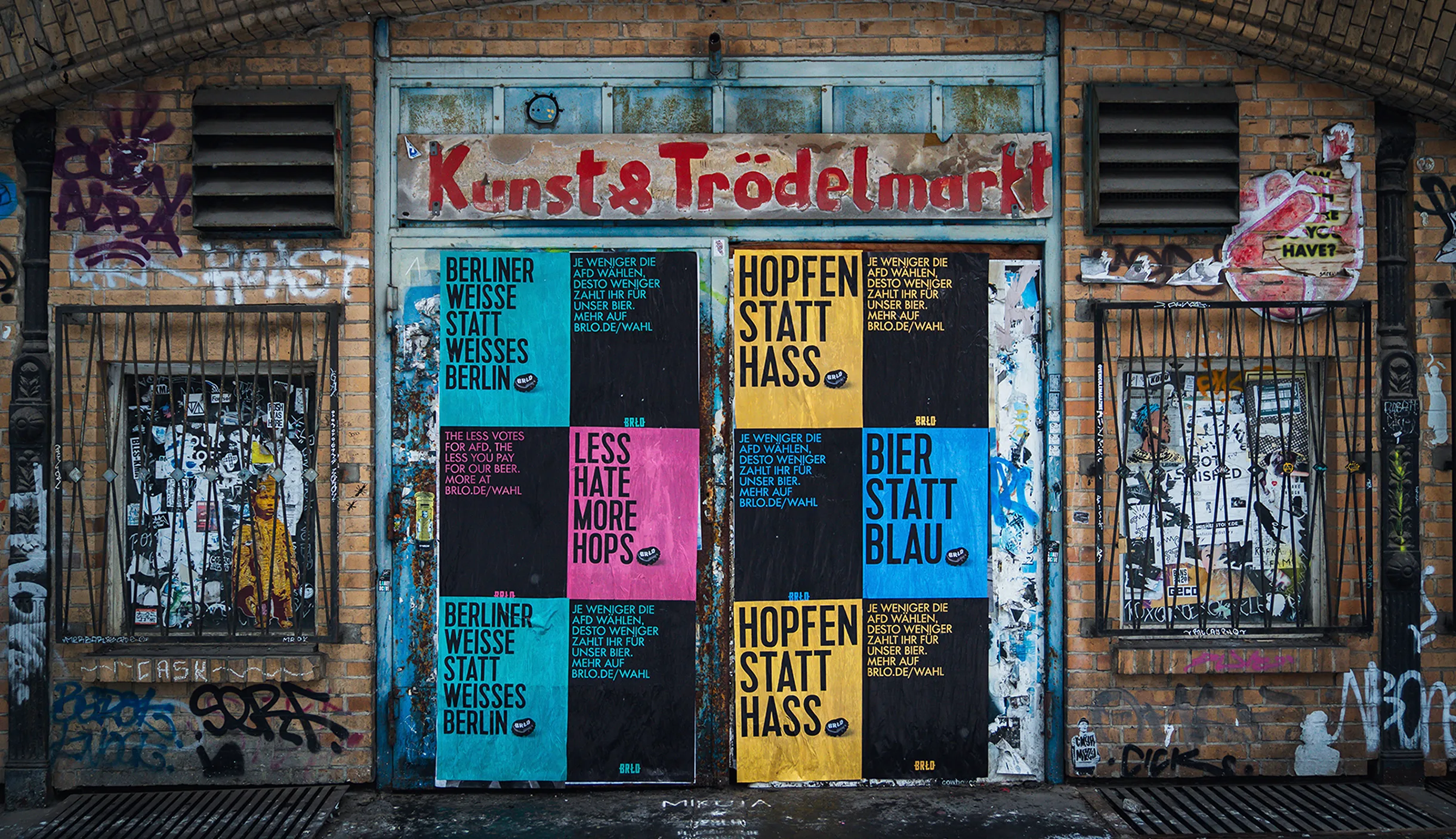 A graffiti-covered brick wall features a closed metal gate with colorful posters in German and English, advertising drinks and promoting positivity. The sign above the gate reads Kunst & Trödelmarkt.