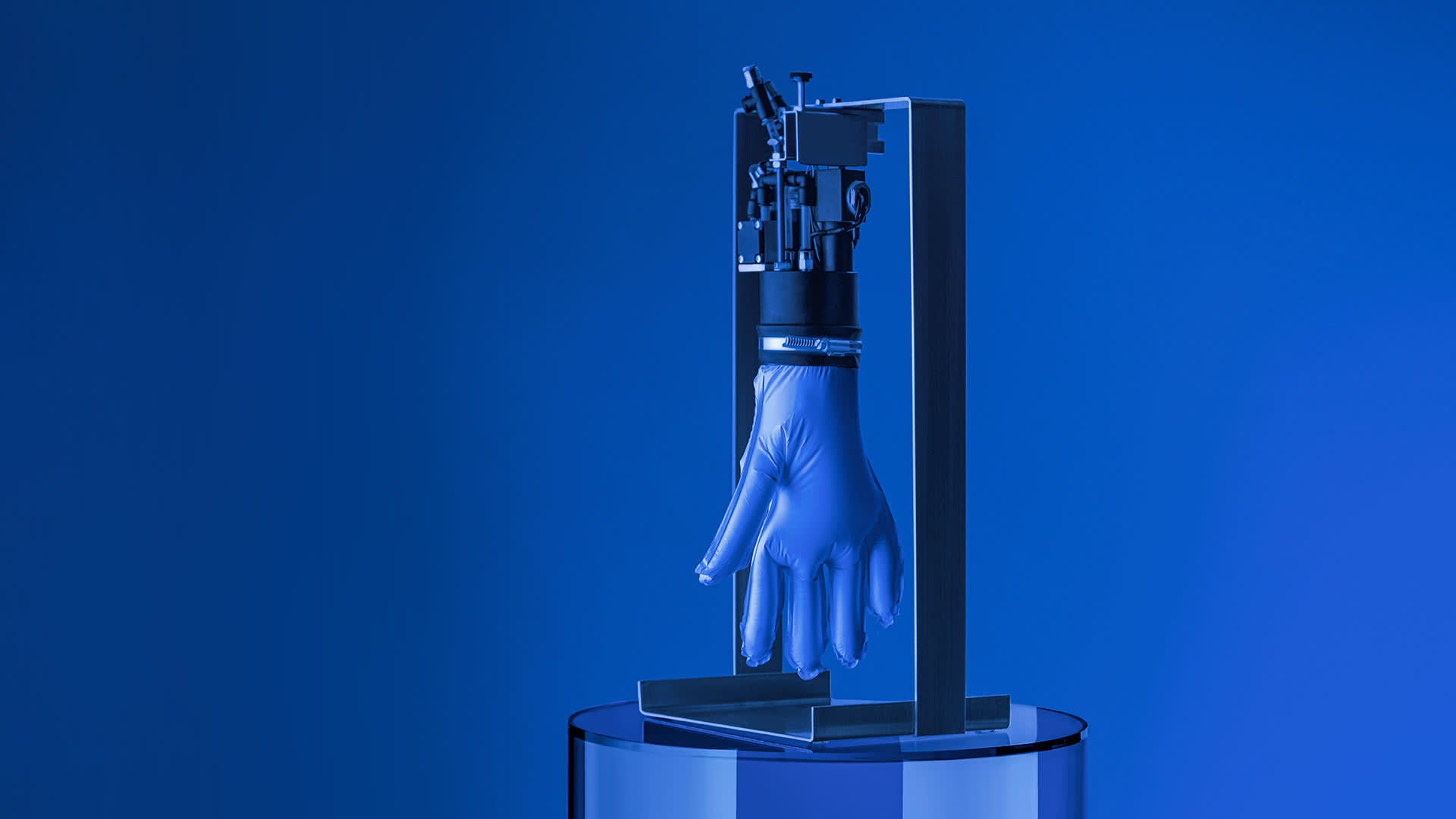 A robotic device holds an inflated blue latex glove upright, with the glove resembling a hand, against a blue background. The setup is positioned on a round, reflective surface.