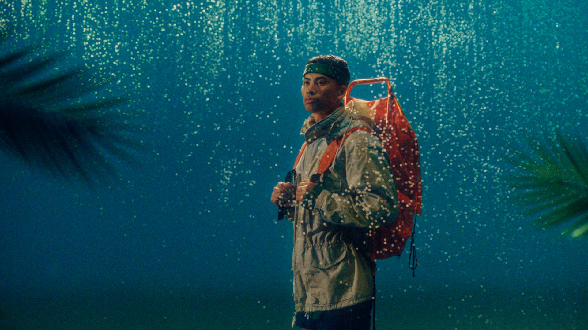 A person wearing a rain jacket and backpack stands in falling rain, illuminated against a blue-green background with blurred plant leaves in the foreground.