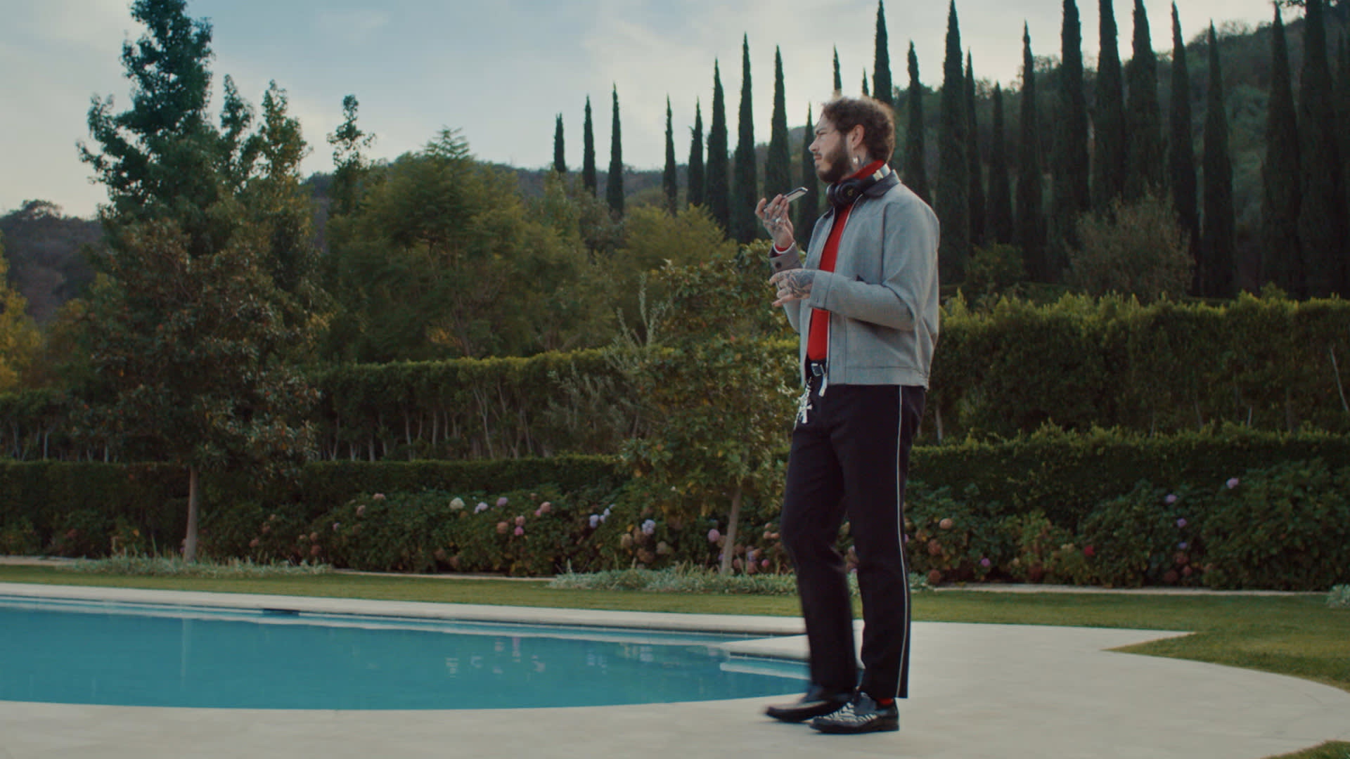A man wearing a gray jacket and dark pants stands by a swimming pool holding a drink, with trees, bushes, and tall cypress trees in the background.