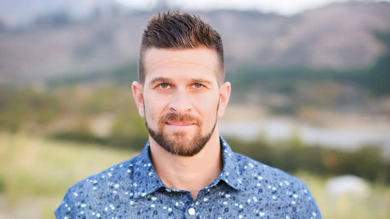 A man with short brown hair and a beard, wearing a blue patterned button-up shirt, stands outdoors with blurred greenery and mountains in the background.