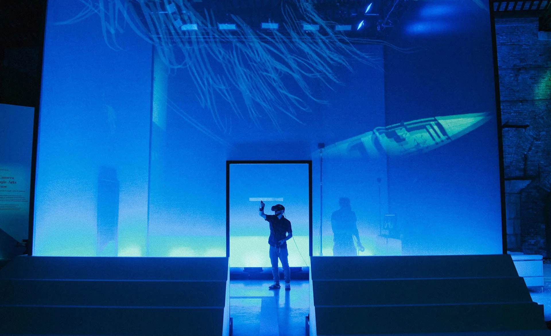 A person stands in a doorway within a blue-lit room, reaching up towards a suspended object. Abstract light patterns and a boat image are projected on the walls, creating a futuristic, immersive environment.