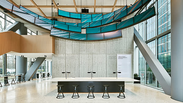 Modern, open lobby space with a long white table and black stools, tall windows, high ceiling, and suspended turquoise panels as decorative elements. Natural light fills the room, creating a bright, airy atmosphere.