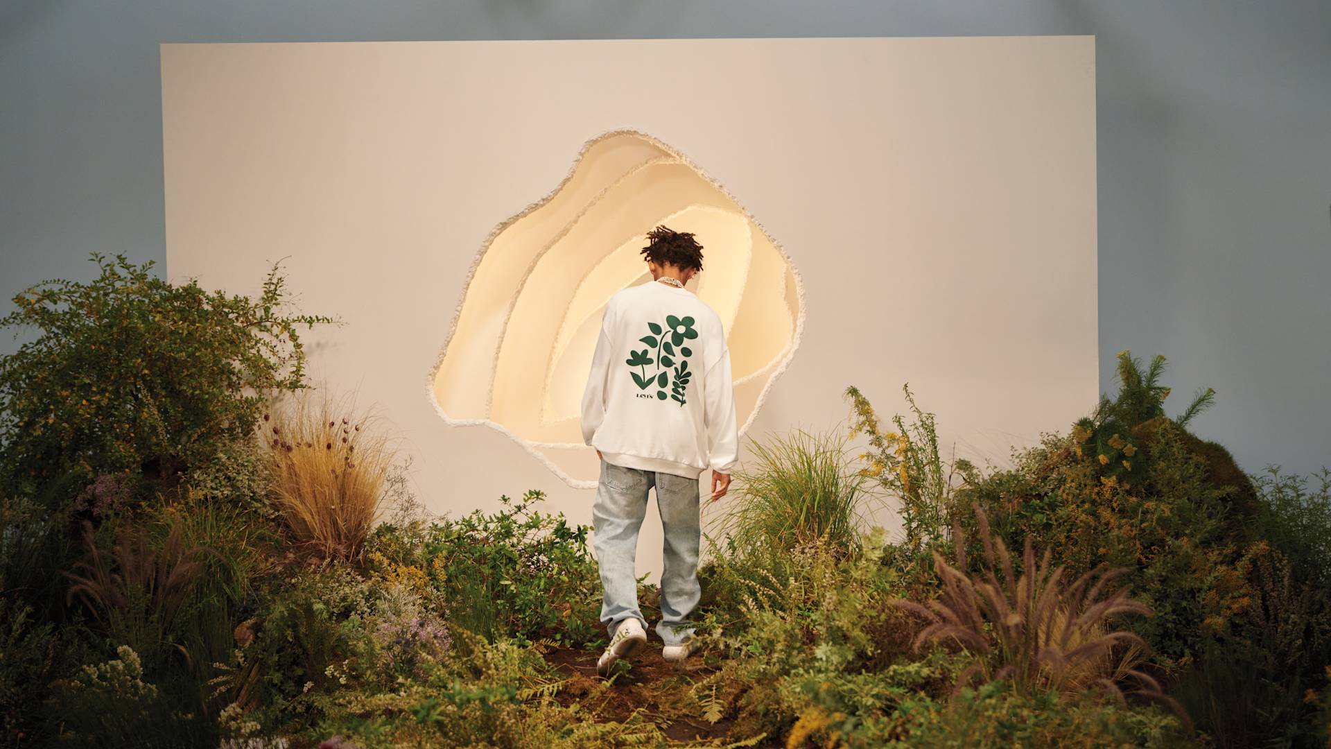 A person wearing a white jacket with a green leaf pattern on the back walks toward a large, irregular opening in a white wall, surrounded by lush greenery and plants.