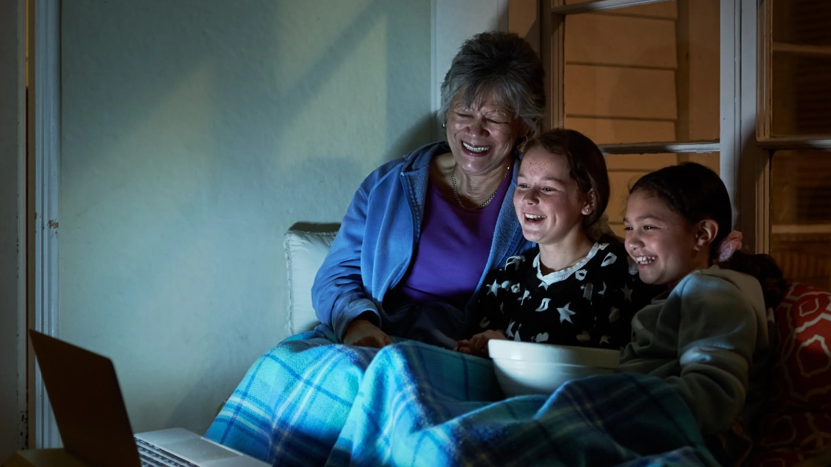 An older woman and two children sit together under a blanket, smiling and watching a laptop screen. They appear to be enjoying a movie, with a bowl of snacks on their laps, in a cozy, dimly lit room.