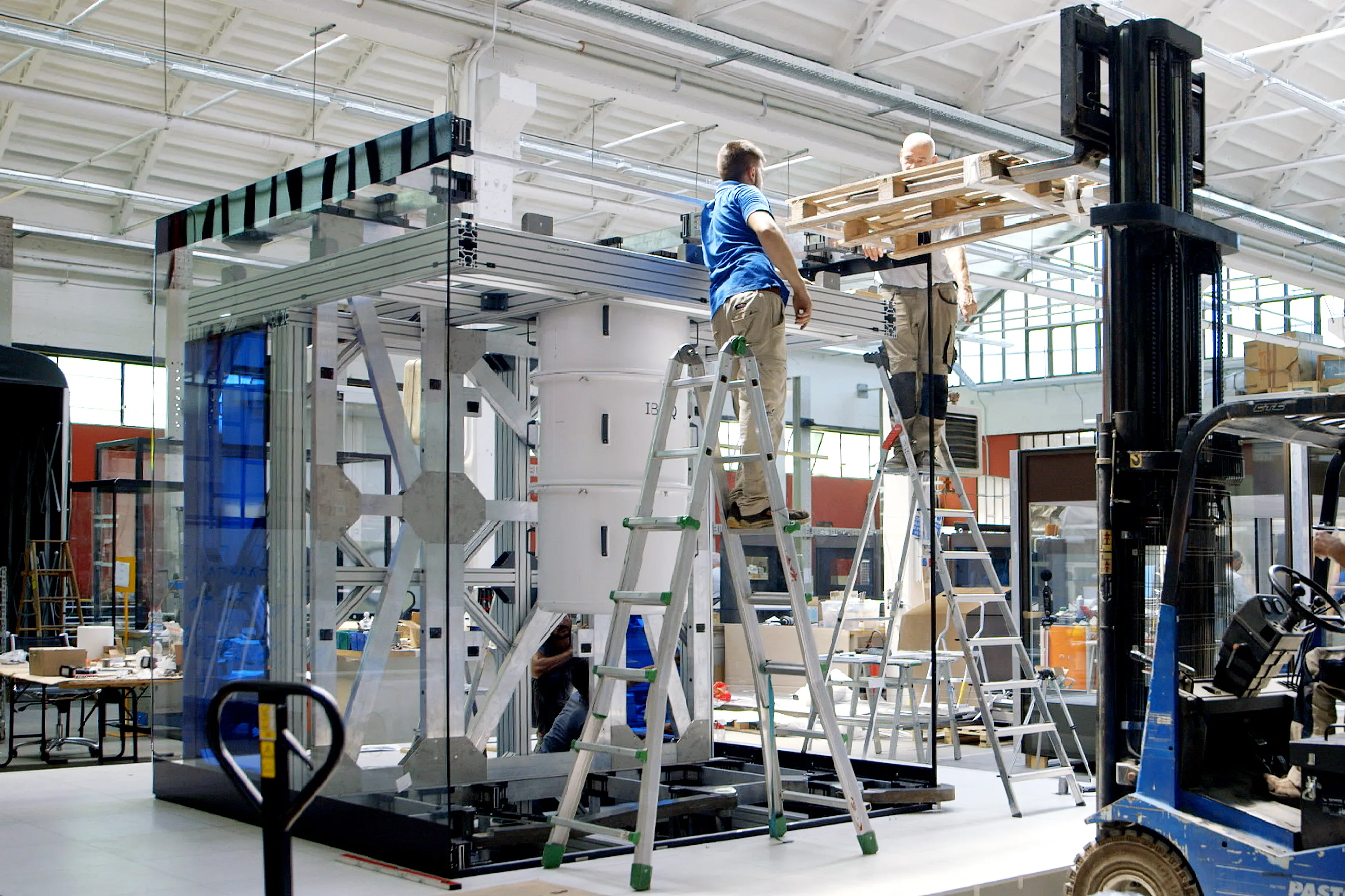 Two workers on ladders assemble or repair a large glass and metal structure indoors, using a forklift to lift materials. The industrial setting includes various tools and machinery in the background.